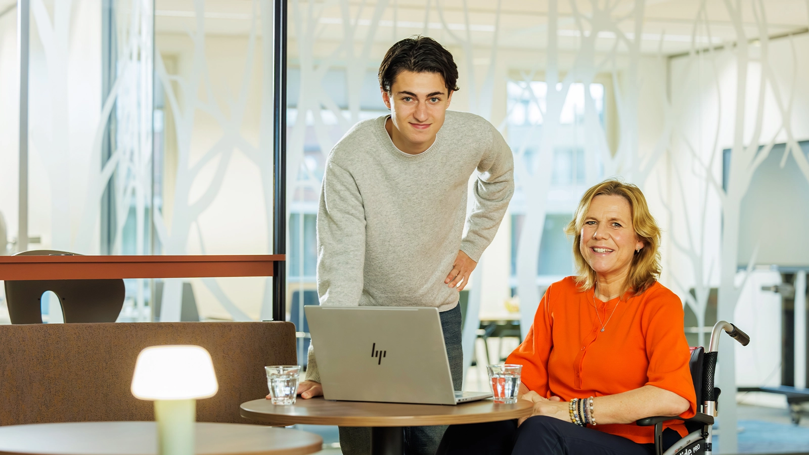 Decoratieve foto van een mannelijke collega en vrouwelijke collega in rolstoel van de Regionale Belasting groep, welke direct in de camera kijken.